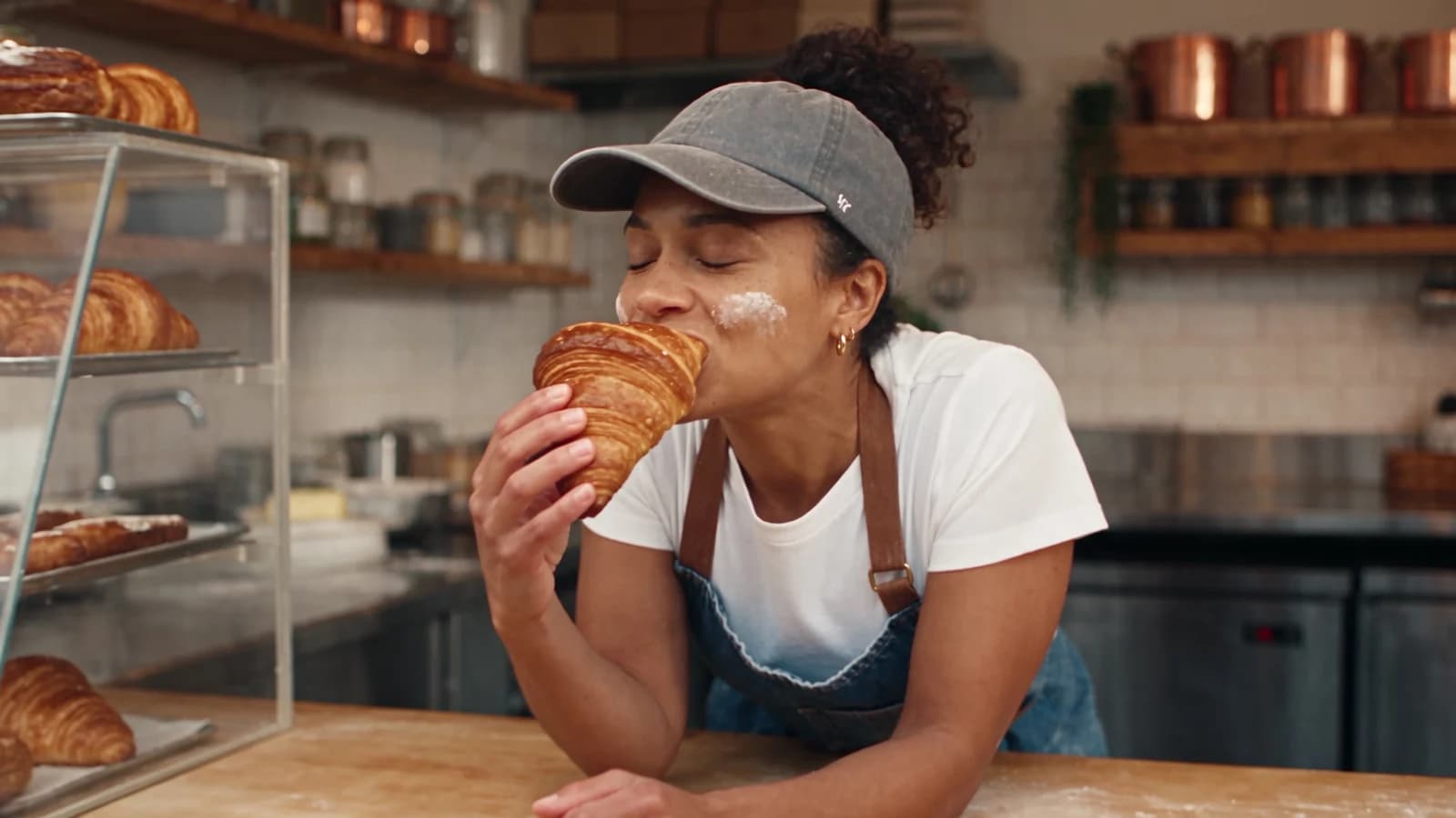 A baker in a denim apron and cap bites into a croissant behind the counter of a small bakery, a frame from the AI film